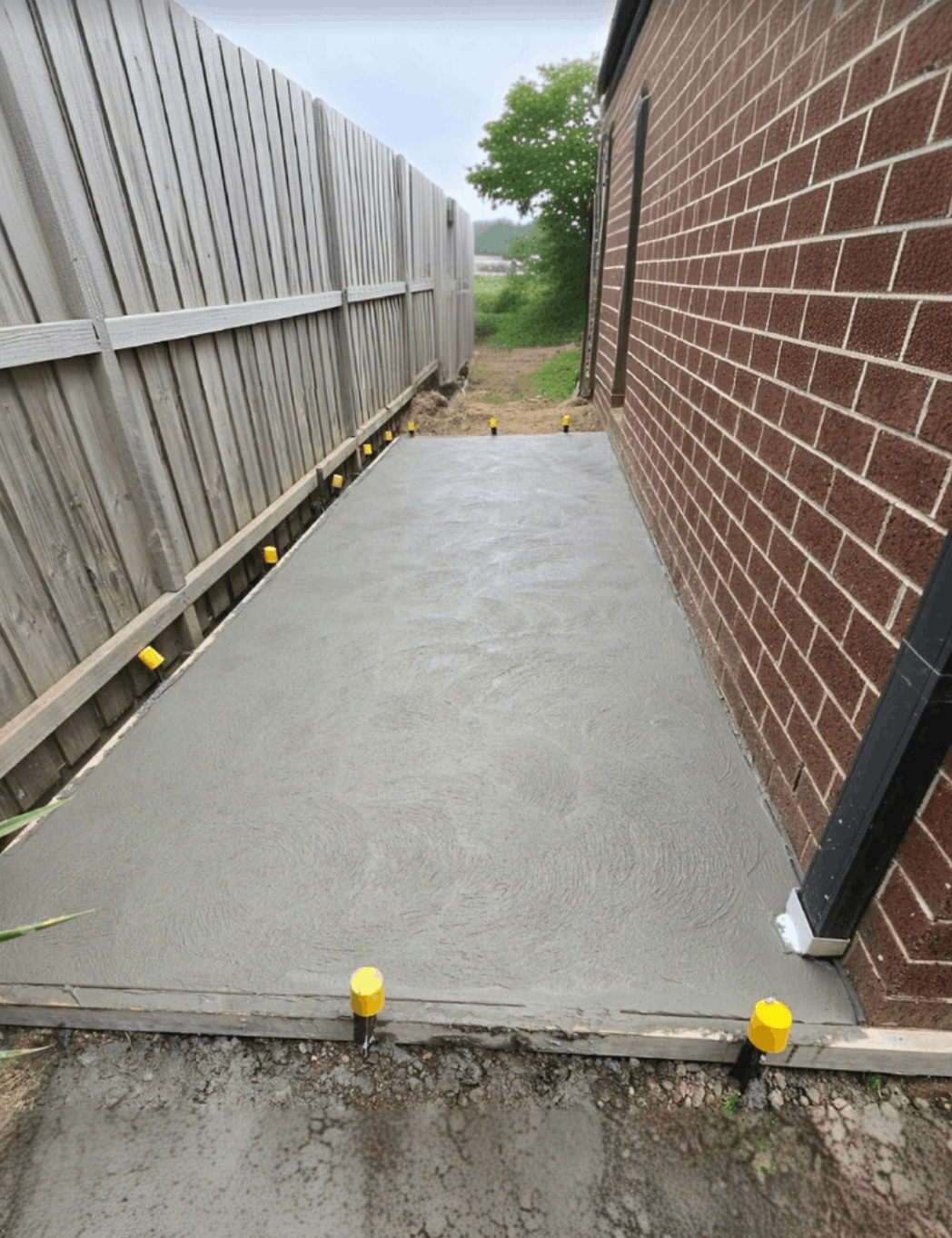 Freshly poured concrete pathway between fence and brick wall